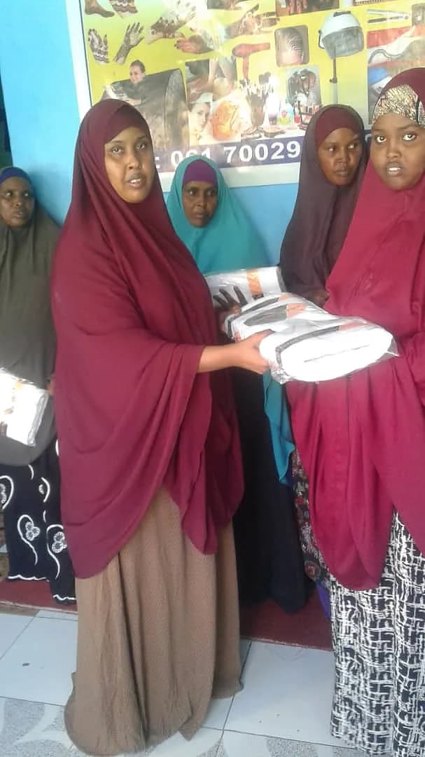 One woman hands wrapped white packages to another, with trainees standing behind them