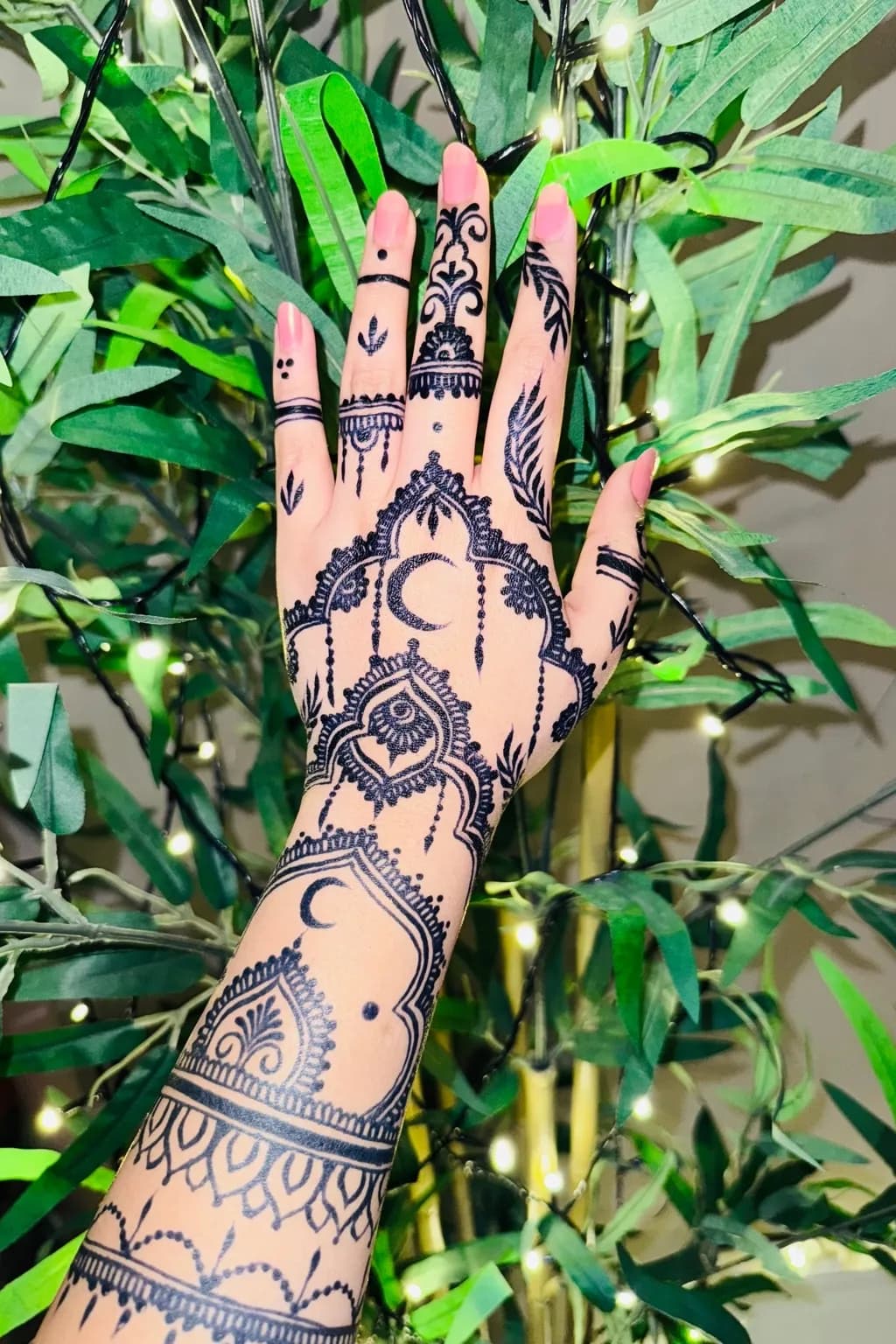 A hand and forearm decorated with fine henna patterns, in front of green leaves